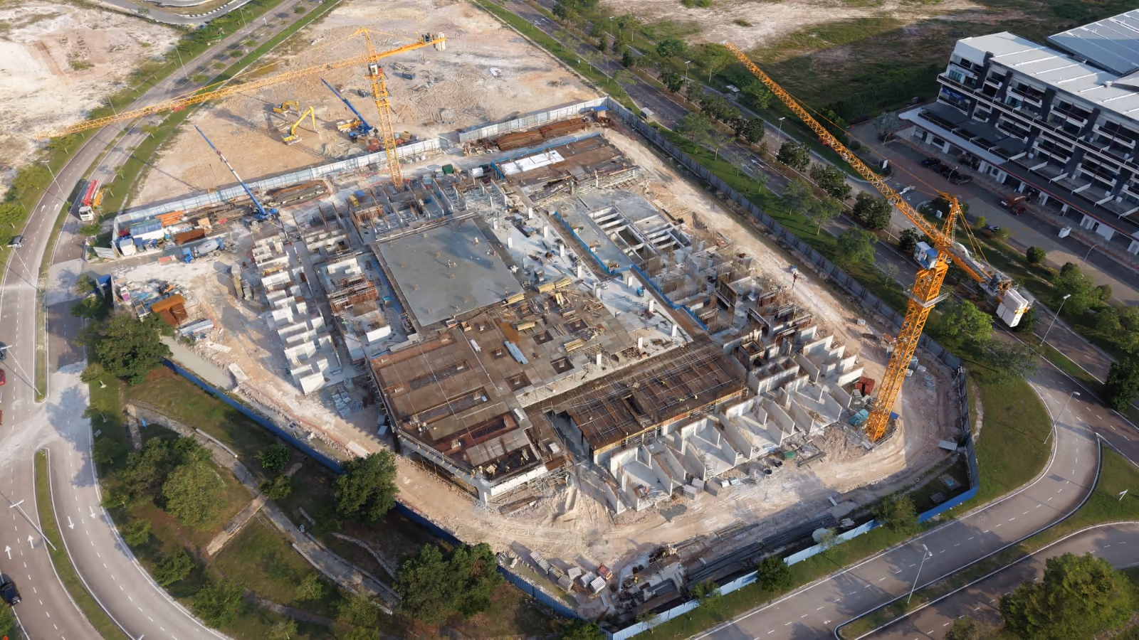 Aerial view of the Bodaiju Residences site in Medini, Iskandar Puteri, in June 2026. Two yellow tower cranes stand over the podium level, where the ground slab has been poured and shear walls and exposed reinforcement are going up behind blue site hoarding, with completed shophouses and cleared land on the surrounding roads.